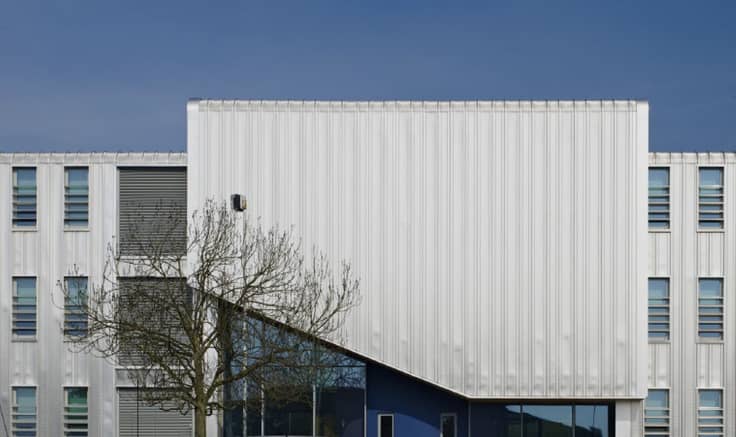 Stylish, simple photograph of the side of a large, white office block with a leaf-less tree in front of it and clear blue sky behind.