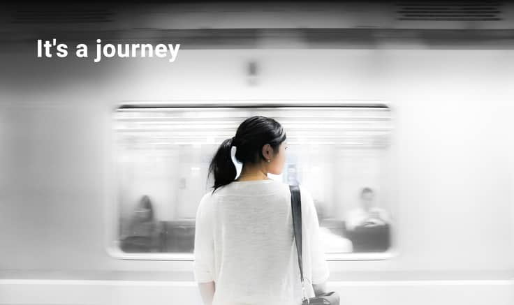 Back view of woman with long, black hair and white shirt, stood in front of a blurred moving train side.